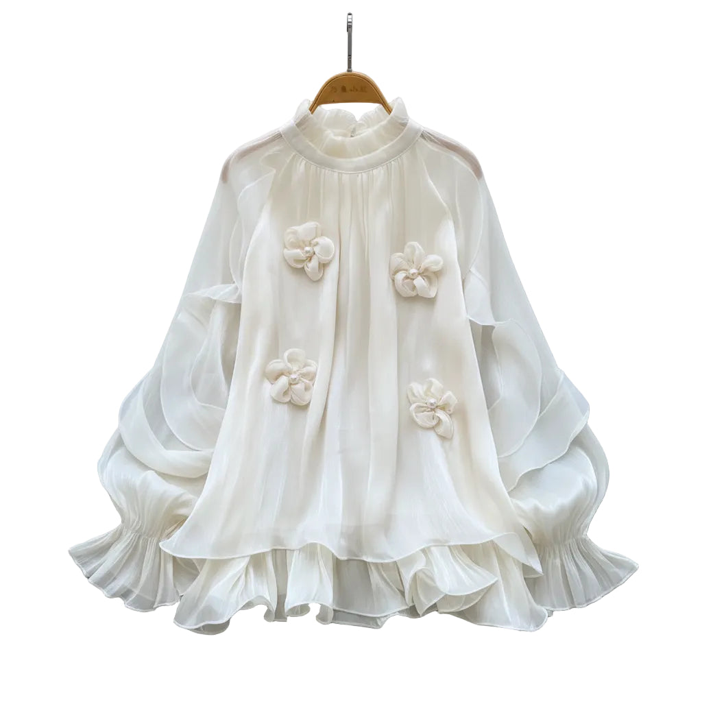 A cream-colored long-sleeve blouse with 3D floral appliqués and a ruffled hem, displayed on a wooden hanger over a white faux fur rug with decorative flowers and a straw hat.