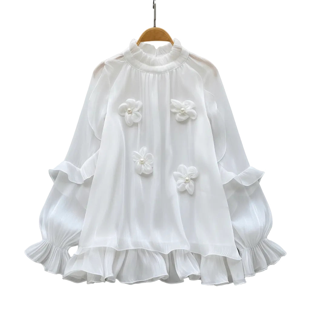 A white long-sleeve blouse with ruffled layers and three 3D floral appliqués, displayed on a wooden hanger on a white faux fur surface with decorative white roses and a straw hat.