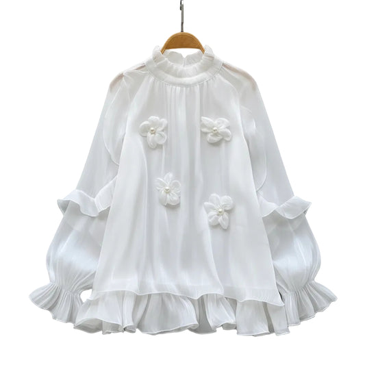 A white long-sleeve blouse with ruffled layers and three 3D floral appliqués, displayed on a wooden hanger on a white faux fur surface with decorative white roses and a straw hat.