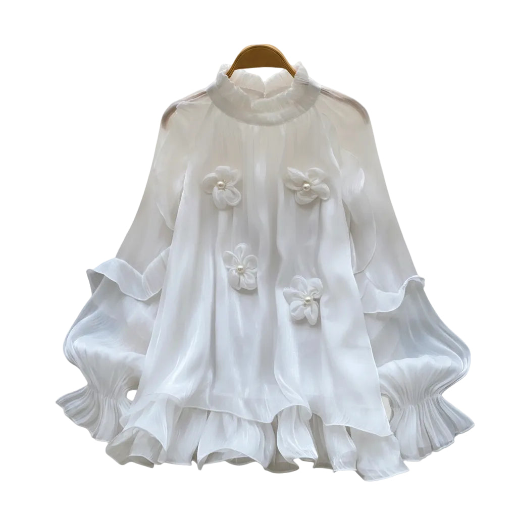 A white long-sleeve blouse with ruffled layers and three 3D floral appliqués, displayed on a wooden hanger on a white faux fur surface with decorative white roses and a straw hat.