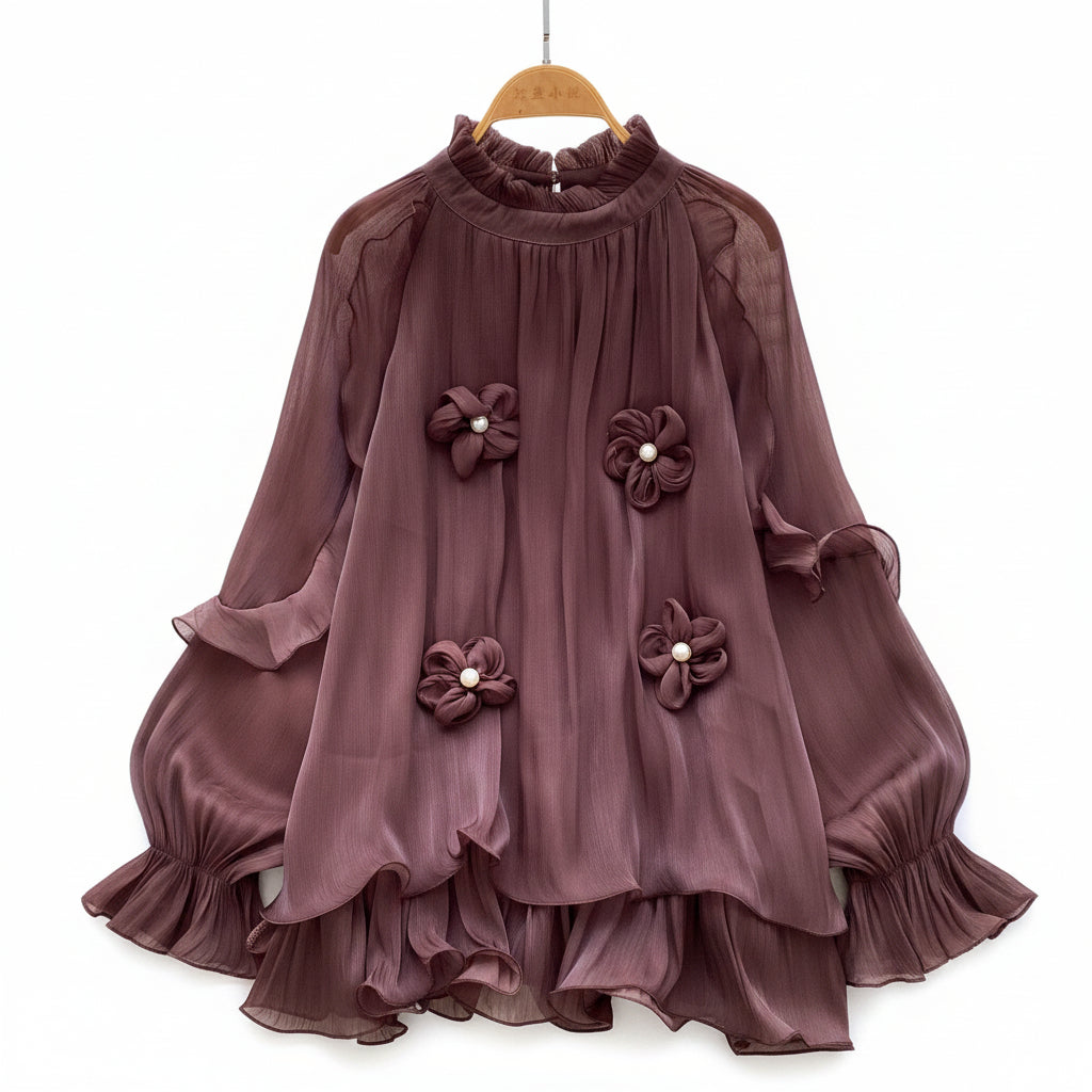 A dark brown long-sleeve blouse with ruffled layers and three 3D flower appliqués on the front, displayed on a hanger over a white faux fur rug with white roses and a straw hat.