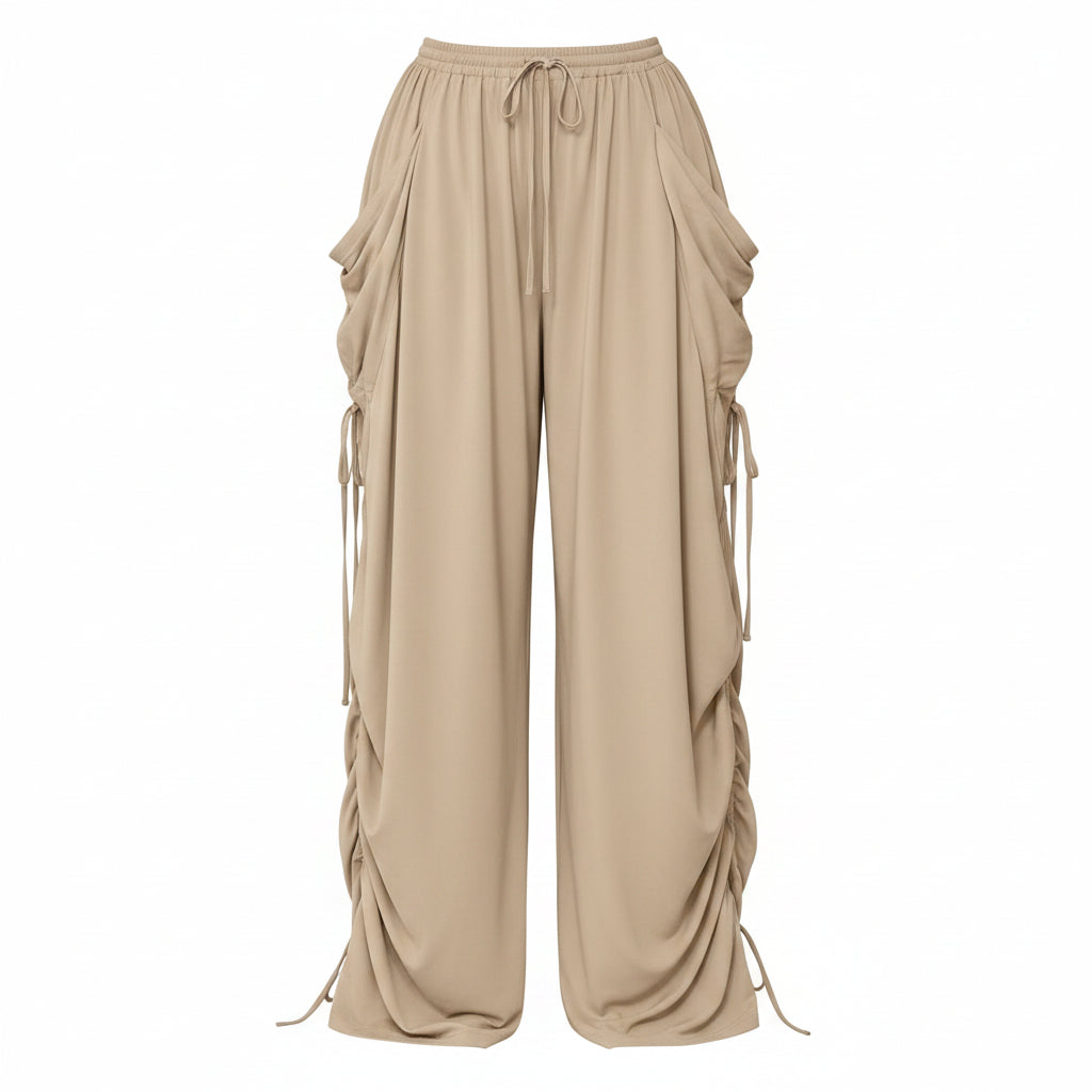 A person wearing loose beige wide-leg drawstring pants with gathered side details and ruched hems, holding a small black handbag. The photo is taken indoors against a plain light wall and gray floor, with 'Plus Size' text in the top-right corner.