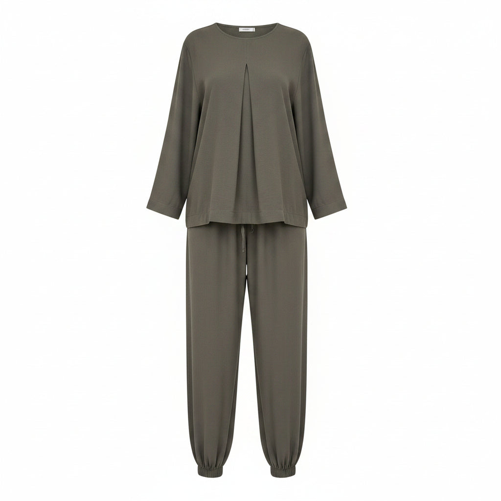 A woman modeling a matching two-piece lounge set in an olive green color, featuring a loose long-sleeve top and matching wide-leg pants with elastic cuffs. She is standing in front of a white closet shelving unit filled with folded white shirts and shoes.