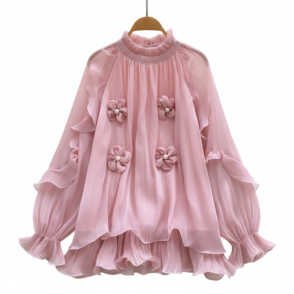 A pink long-sleeve blouse with layered ruffles and three 3D fabric flower embellishments on the front, displayed on a wooden hanger over a white wire rack with decorative props including white roses, a straw hat, and a white purse.