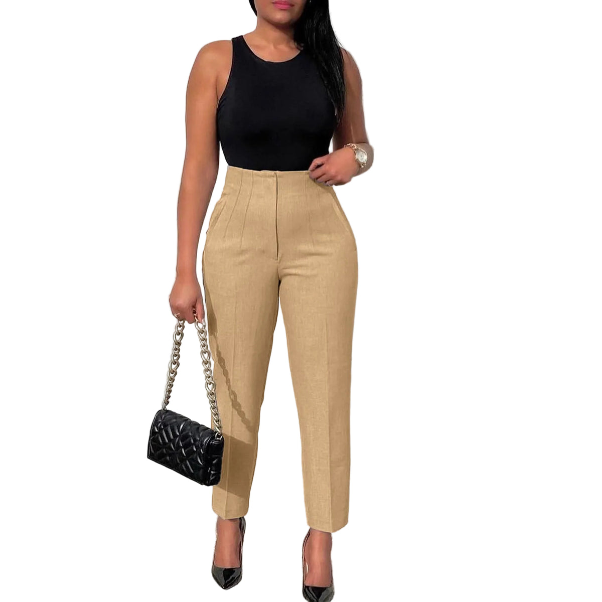 Woman wearing a black top and beige pants holding a black handbag against a light gray wall.