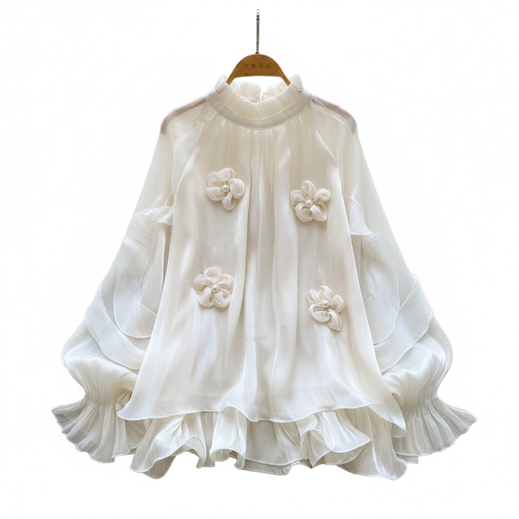 A cream-colored long-sleeve blouse with 3D floral appliqués and a ruffled hem, displayed on a wooden hanger over a white faux fur rug with decorative flowers and a straw hat.