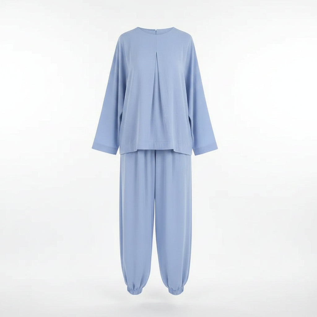 A woman modeling a light blue two-piece lounge set consisting of a long-sleeve top and matching wide-leg pants, styled with a small white handbag and sunglasses in front of a white closet filled with folded clothes and shoes.