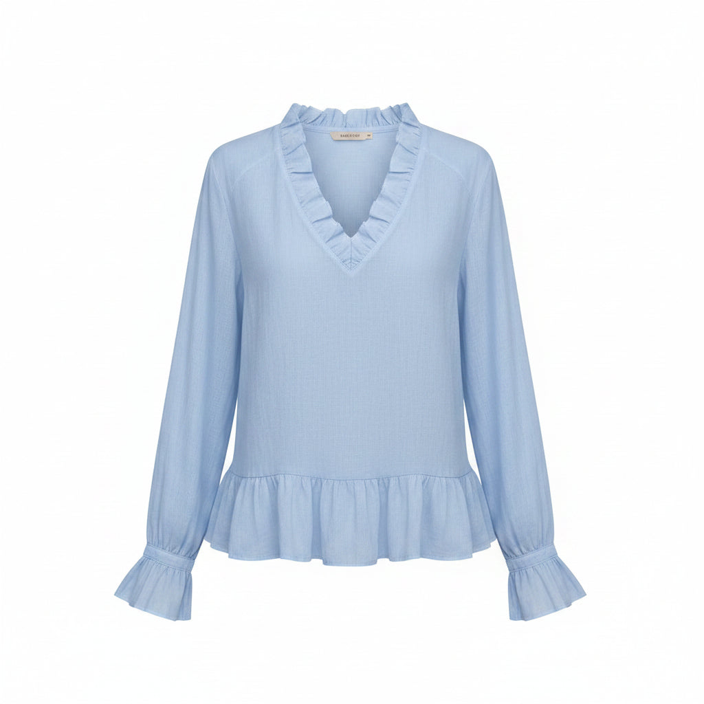 A woman wearing a light blue long-sleeve blouse with a ruffled V-neckline and ruffled cuffs, paired with off-white pants, against a neutral gray background.