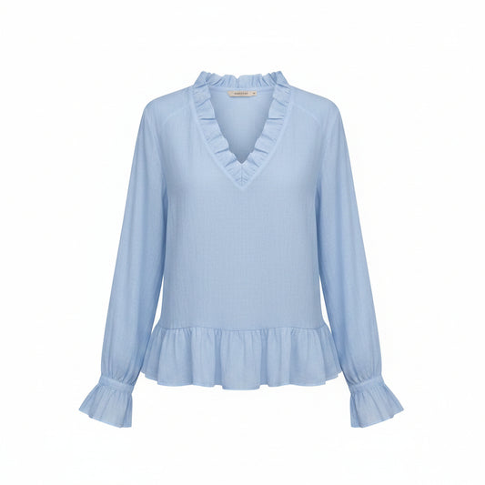 A woman wearing a light blue long-sleeve blouse with a ruffled V-neckline and ruffled cuffs, paired with off-white pants, against a neutral gray background.