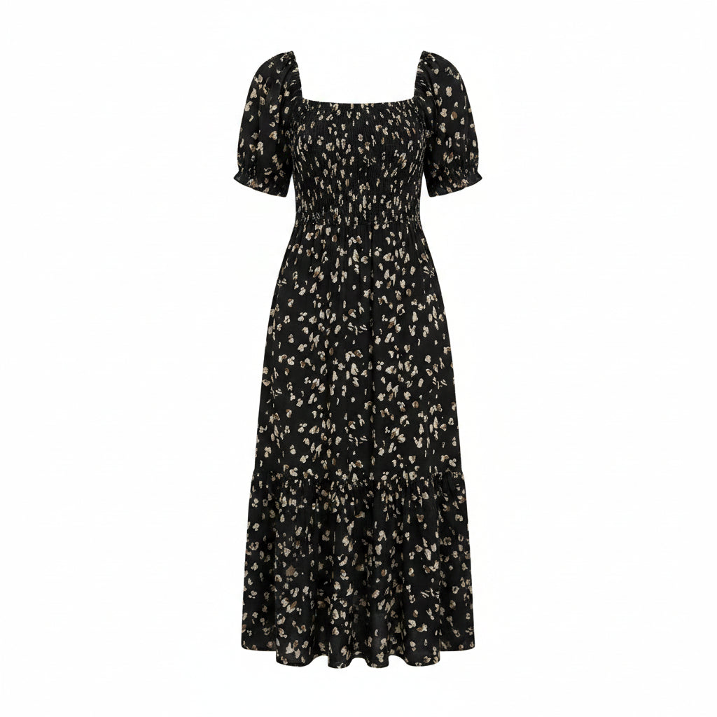 A woman wearing a black midi dress with a small beige floral print, holding a woven straw bag. The dress features a smocked bodice, short flutter sleeves, and a tiered skirt, shown in a lifestyle setting against a white wall and green plants.