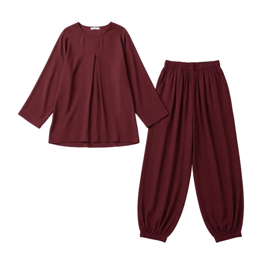 A woman modeling a matching burgundy two-piece lounge set consisting of a long-sleeve top and wide-leg pants, styled with a white handbag and sunglasses in front of a white closet filled with folded white shirts and shoes.