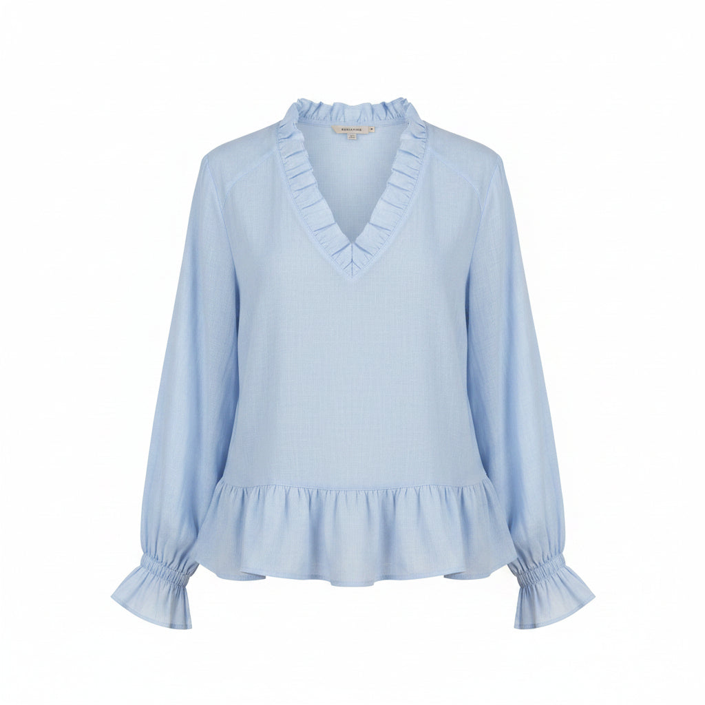 A woman wearing a light blue long-sleeve blouse with a ruffled V-neckline, ruffled cuffs, and a ruffled hem, paired with cream pants against a neutral gray background.